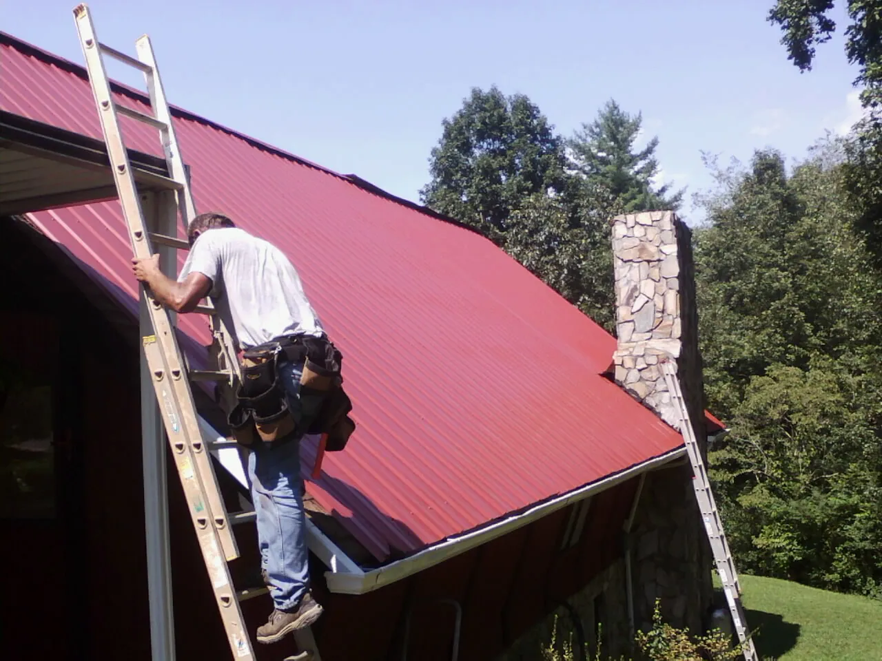 Roofer on a ladder beside a red metal roof and stone chimney — Soffit & Fascia Repair by Roofing Brothers in Lake Lillian